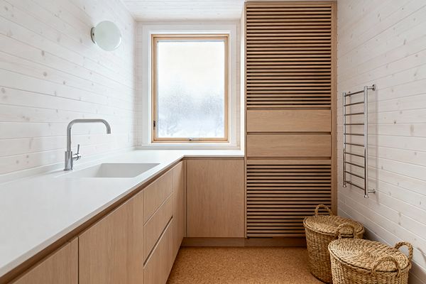 Minimal Scandinavian laundry room with whitewashed wood panel walls and ceiling, a long white countertop with integrated rectangular sink, and flat-front light oak cabinets along the left and back walls. A frosted wood-framed window centers the space, next to a tall slatted oak cabinet, stainless towel rail, and two lidded woven baskets on warm cork flooring, lit by a round opal wall sconce.