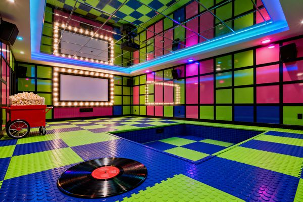 A playful home media room features a large screen framed by marquee bulbs, glossy wall panels in lime green and hot pink set within a black grid, and a checkerboard floor of blue and green raised-stud tiles. A mirrored ceiling, recessed blue-tiled pit, colored LED cove lighting, wall speakers, a ceiling projector, a red popcorn cart, and a vinyl record on the floor complete the graphic Pop Art look.