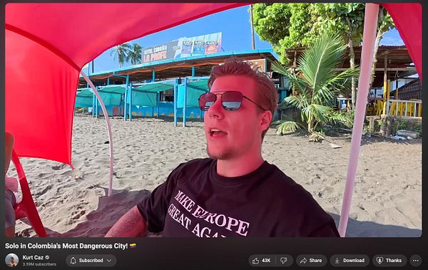 A man wearing a black t-shirt with a slogan sits under a red beach umbrella on a sandy beach.