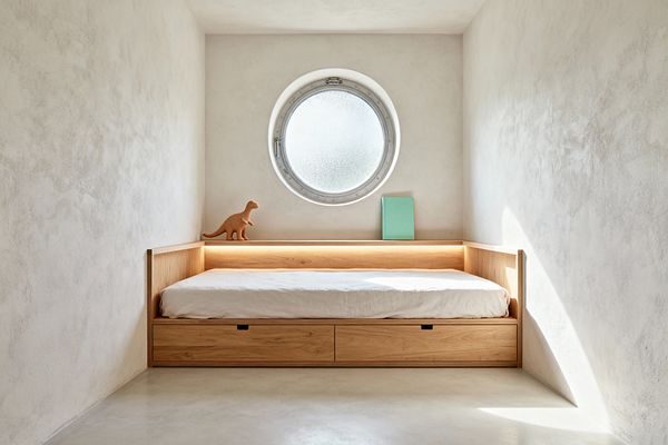 An intimate minimalist kids’ room features a built-in light oak daybed with two drawer fronts and a wraparound low back that forms a shelf. Centered above, a round frosted-glass porthole window diffuses daylight onto warm white plaster walls and a pale concrete floor, while concealed LED lighting glows along the shelf; a terracotta dinosaur toy and a mint green book add small accents.