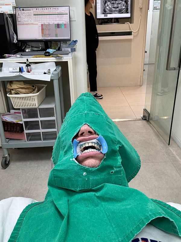 A person is lying in a dental chair with a green dental bib and a mouth guard, while a dental assistant stands in the background.
