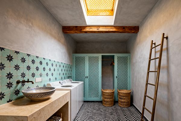 A Moroccan-style laundry room with soft gray plaster walls features a mint zellige tile backsplash patterned with navy starbursts. A beige travertine console holds a blue-patterned ceramic vessel sink and a wall-mounted brass faucet. A white washer and dryer sit below the tiles. Opposite, mint lattice-front cabinets and an open hanging nook stand beside two woven rattan hampers. A brass wall ladder, black-and-white chevron cement floor, timber beam, and a skylight with a perforated screen complete the space.