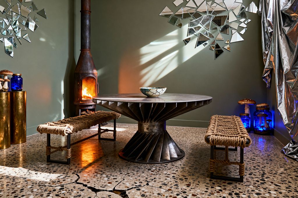 A round, weathered-steel dining table with a turbine pedestal sits between two woven jute-rope benches on a speckled terrazzo floor marked by visible cracks. A small, rusted stove glows at left against sage-green walls near a brass pedestal with a cobalt jar. Shattered mirror tiles scatter across the wall, while silver mylar drapery and blue-lit glass jars with sculptural mushrooms create a survivalist mood as sunlight and firelight cast sharp reflections.