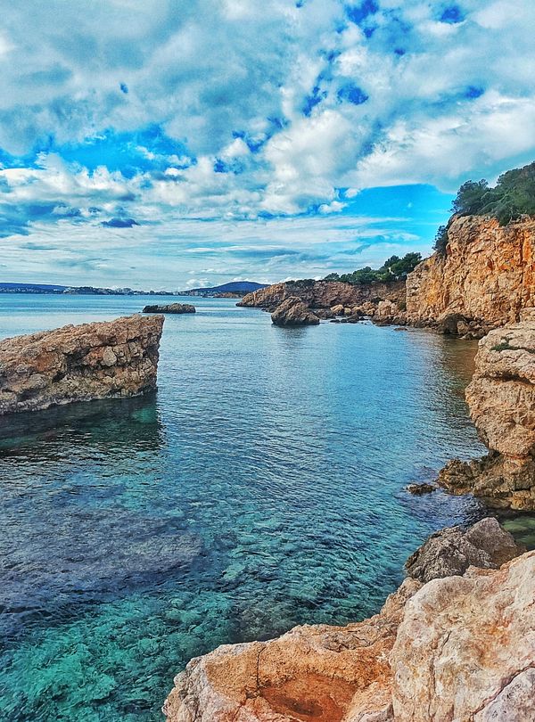 A scenic coastal view featuring rocky cliffs and clear blue waters under a partly cloudy sky.