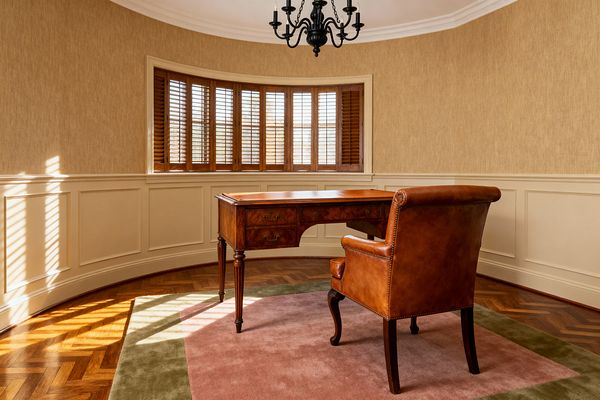 A traditional home office features a curved bay window with wood shutters casting light over a herringbone wood floor. Centered in the room is a mahogany writing desk with a leather inlay and brass pulls, paired with a caramel leather armchair trimmed with nailheads. Cream paneled wainscoting and crown molding wrap the space, above which beige textured wallcovering meets a white ceiling and a black wrought-iron candelabra chandelier.