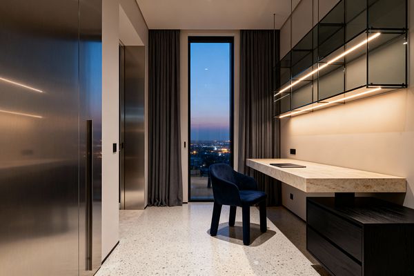 A narrow contemporary home office featuring a floating travertine desk anchored to a black wood drawer unit, with smoked-glass wall cabinets above lit by warm linear LEDs. A deep navy velvet chair sits on light terrazzo flooring. One wall is clad in brushed stainless-steel panels; the opposite wall is soft beige. Floor-to-ceiling charcoal curtains frame a tall window overlooking a city skyline at dusk, with minimal black outlets and slim hardware punctuating the space.