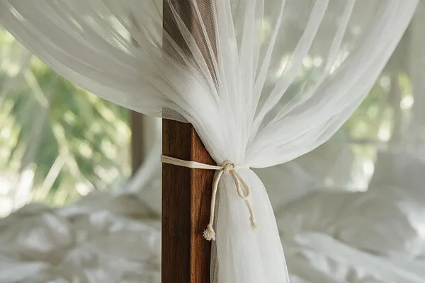 A close-up of a tropical bed canopy with airy white sheer fabric gathered around a dark wooden post and tied with a thin cord. Soft bedding and blurred palm greenery in the background create a relaxed, breezy resort feel.