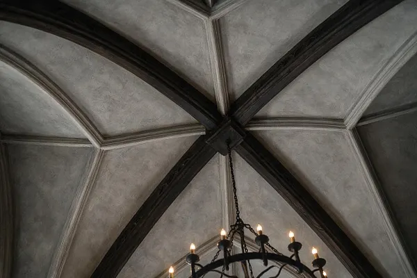 An upward view of a Gothic-style vaulted ceiling with pale plaster panels framed by dark timber ribs forming pointed geometric sections. A wrought iron chandelier with candle-style bulbs hangs from the center, adding a dramatic historic feel.