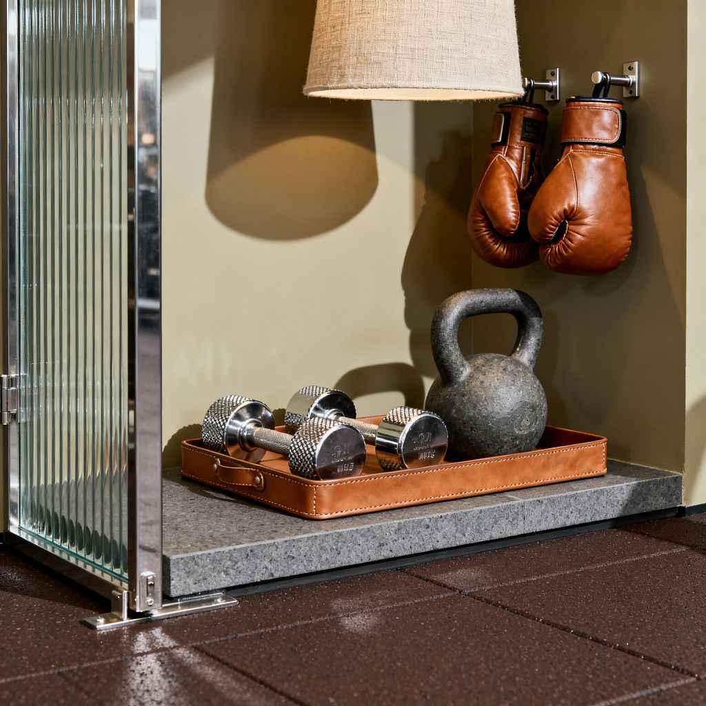A close-up of a transitional home gym vignette: a fluted glass divider with polished chrome frame sits beside a muted olive wall lit by a linen-shaded sconce. Cognac leather boxing gloves hang from metal hooks above a gray stone ledge, where a leather tray organizes two chrome dumbbells and a cast-iron kettlebell; dark brown rubber floor tiles complete the space.