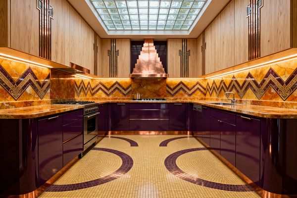 A U-shaped Art Deco kitchen features glossy aubergine base cabinets with copper trim, amber onyx countertops, and a matching backsplash patterned with purple-and-gold chevrons. Centered by a stepped copper range hood, light oak upper cabinets carry vertical inlays; warm undercabinet LEDs highlight the stone, a cream-and-plum mosaic floor sweeps in arcs, a glass-block skylight brings daylight, and a stainless range and single-bowl sink complete the space.