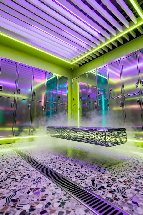 A futuristic mudroom lined with reflective stainless-steel lockers glows under purple and lime LED lighting. A perforated metal bench appears to float above a terrazzo floor with a long linear drain, while mist and underlighting add a dramatic effect. Overhead, white slatted ceiling baffles integrate light strips, and neon-lime walls bounce color across the metal surfaces. The space is empty of people and highly clean-lined and industrial.