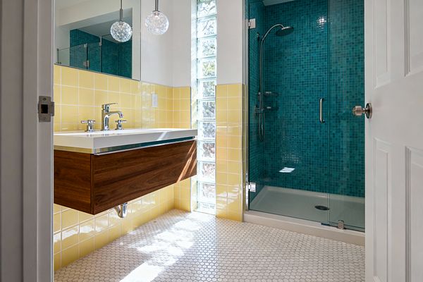 A mid-century modern bathroom with a floating walnut vanity and white countertop beneath mirrored cabinets and two bubble-glass pendants. Butter-yellow square tiles wrap the lower walls, a vertical column of glass blocks brings in daylight, and a glass-enclosed shower lined in teal mosaic tile anchors the back, while a white penny tile floor and polished chrome fixtures complete the bright, clean look.