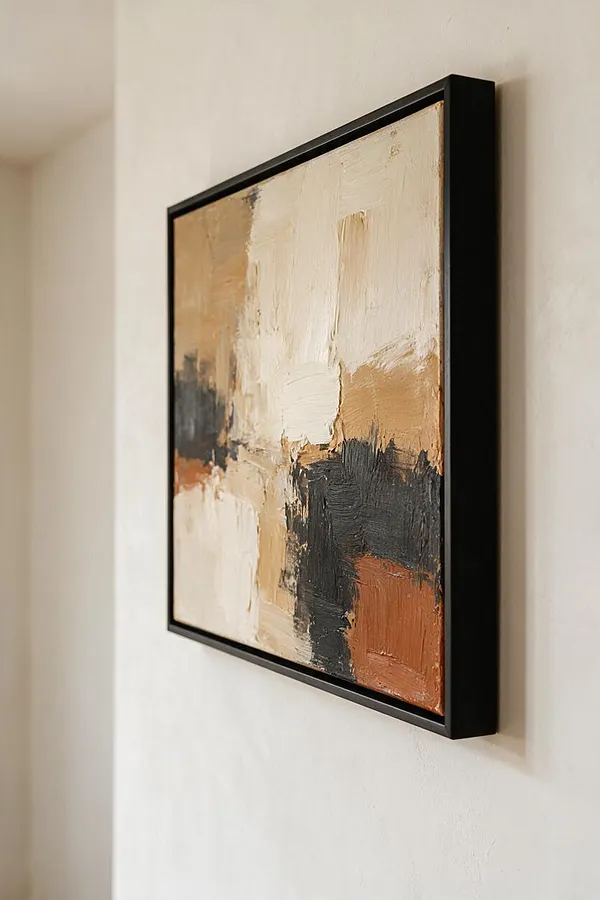 A framed abstract painting hangs on a light beige wall, featuring layered brushstrokes in cream, taupe, charcoal, and rust. The slim black frame gives the artwork a crisp modern edge.