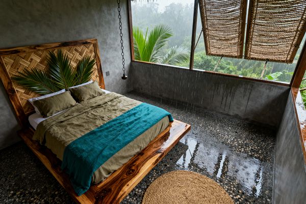 An open-air tropical bedroom features a live-edge wood platform bed and a woven rattan headboard accented with large palm fronds. Olive linen bedding is layered with a teal throw. Gray troweled concrete walls open to a jungle view through a wide, glassless window where rain falls onto a pebble-set floor. Rope-woven roll-up shades hang above the opening, a chain-hung pendant sits beside the bed, and a round jute rug softens the wet stone surface.