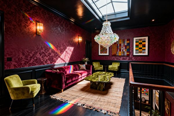A dramatic maximalist living room with burgundy damask wallpaper above black wainscoting and a glossy black ceiling punctuated by a large rectangular skylight. A tiered chandelier of translucent shells hangs over a sculpted green onyx coffee table. A magenta velvet sofa and two chartreuse velvet armchairs sit on a fringed jute rug atop dark wood floors. Brass lantern sconces and colorful geometric artwork line the walls, while sunlight through the skylight casts rainbow prisms across the space.