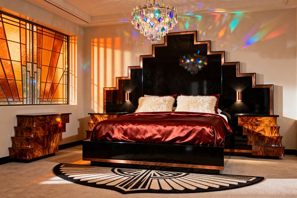 An Art Deco bedroom with a glossy black, stepped ziggurat headboard trimmed in warm metal, flanked by matching stepped nightstands in walnut burl. A bed dressed in deep garnet satin with two white faux-fur pillows sits on a black platform above a fan-motif rug; an amber stained-glass window and a tiered crystal chandelier cast geometric shadows and rainbow reflections onto cream walls and pale carpet.