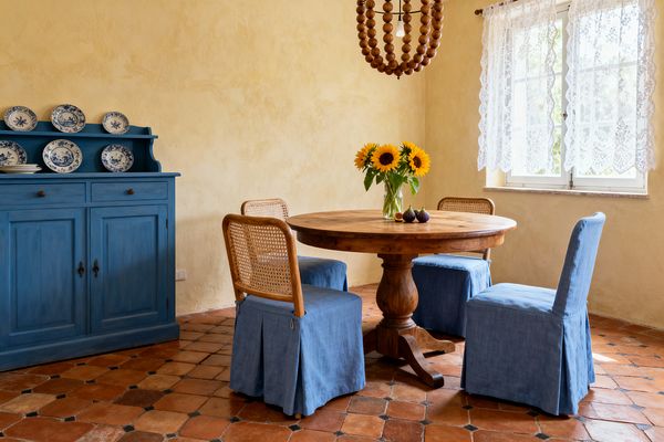 A sunlit French Country dining room features an oak round pedestal table with a vase of sunflowers, surrounded by four chairs: two with woven cane backs and blue slipcovered seats, and two fully slipcovered in blue linen. To the left, a Provence-blue buffet displays blue-and-white plates; terracotta floor tiles, ochre plaster walls, lace window curtains, and a wood-bead chandelier add rustic texture.