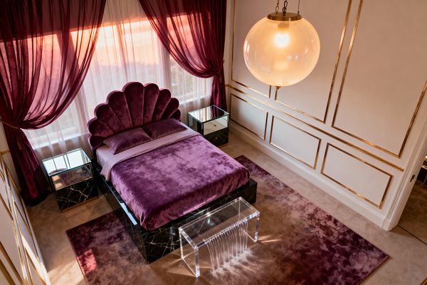 A glamorous bedroom showcases a scalloped, shell-shaped headboard upholstered in deep plum velvet with matching bedding on a black marble plinth base. Symmetrical mirrored nightstands, white walls with gold panel molding, and a brass-hung globe pendant add shine, while burgundy drapery layered over white sheers, a mauve area rug on beige carpet, and a clear acrylic waterfall bench complete the Hollywood Regency look.