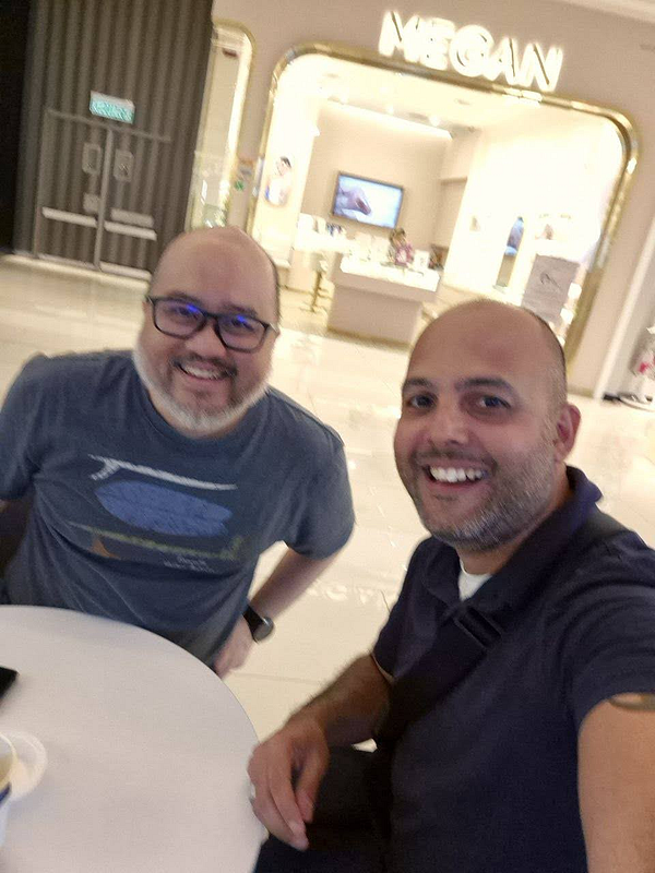 Two men are smiling and posing for a selfie at a café in a shopping area.