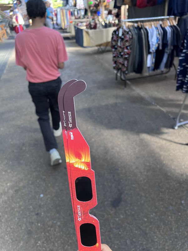 Photo of a crowded outdoor market stall selling solar eclipse glasses with London price tags.