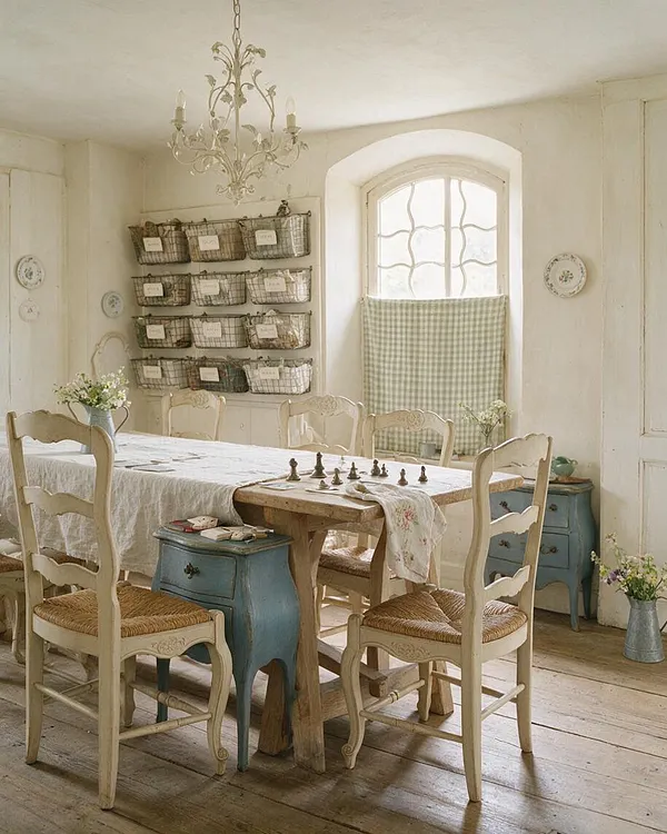 A French Country-style game room with a rustic wooden table surrounded by cream-painted chairs with woven seats, a soft white tablecloth, and a chess set at the center. The room features a wrought-iron chandelier, a large arched window with a gingham curtain, weathered blue side tables, and baskets mounted on the wall for storage.