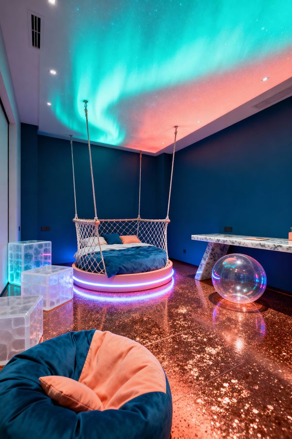 A kids' room with deep teal walls features a round bed suspended from the ceiling by thick rope and wrapped in a net, its circular base rimmed with RGB lights. Above, a neon aurora of cyan and pink glows across the ceiling, reflecting on a copper-glitter epoxy floor. Frosted acrylic cube stools, a marble-look Z-leg desk with underlighting, a clear inflatable ball, and a teal-and-peach velvet bean bag complete the futuristic, playful setting.