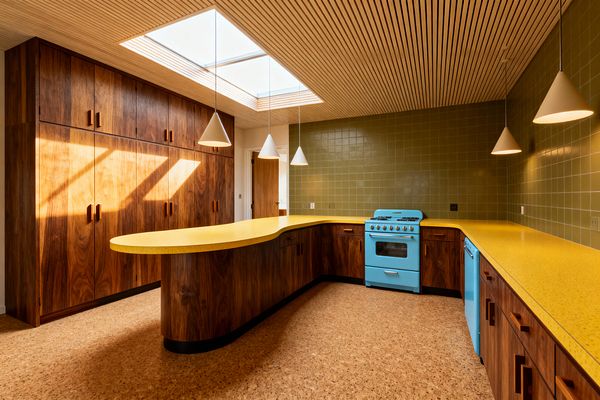 A Mid-Century Modern kitchen features flat-front walnut cabinets with integrated wood pulls and a curved peninsula topped in speckled mustard-yellow laminate. Olive green square tiles line the backsplash, while a retro aqua-blue range and matching dishwasher add color. The space has cork flooring, a slatted light-wood ceiling with a large skylight, and white cone pendant lights that hang over the counters.
