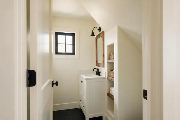 A narrow farmhouse powder room with creamy white walls, a small black-framed window, and a white vanity topped with a light countertop. The room features a rustic wood-framed mirror, black wall sconces and faucet, open built-in shelves with woven baskets and folded towels, and dark floor tiles for contrast.