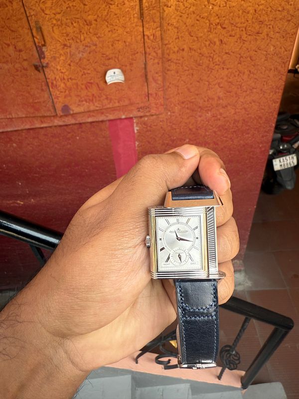 A hand holds a stylish silver watch against a textured orange background.