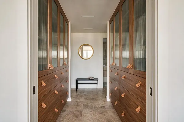 A narrow modern closet hallway with symmetrical built-in wardrobes on both sides, featuring warm wood veneer drawers, copper-toned leather pulls, and upper cabinet doors with reeded glass panels. A round gold-framed mirror and a small black bench sit at the end of the space over large-format stone-look tile flooring.