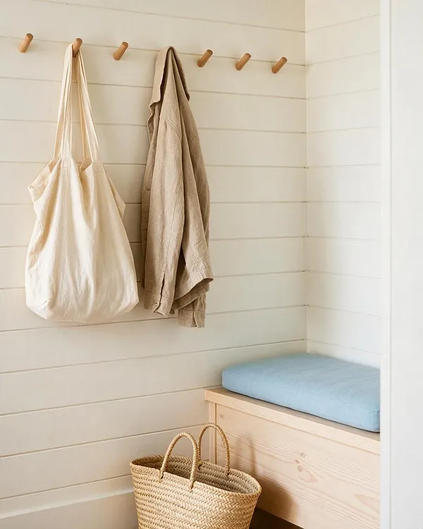 A bright coastal-style mudroom nook with white horizontal shiplap walls, wooden wall pegs holding a cream tote and a beige jacket, a pale wood bench topped with a soft blue cushion, and a woven straw basket on the floor. The palette is airy and natural with relaxed textures.