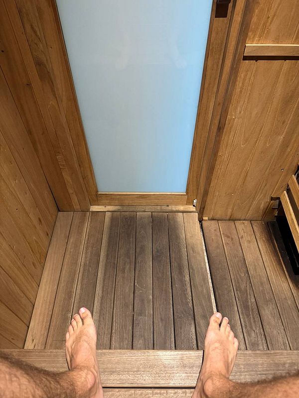 A person's feet are visible as they sit inside a sauna, with wooden flooring and a glass door.