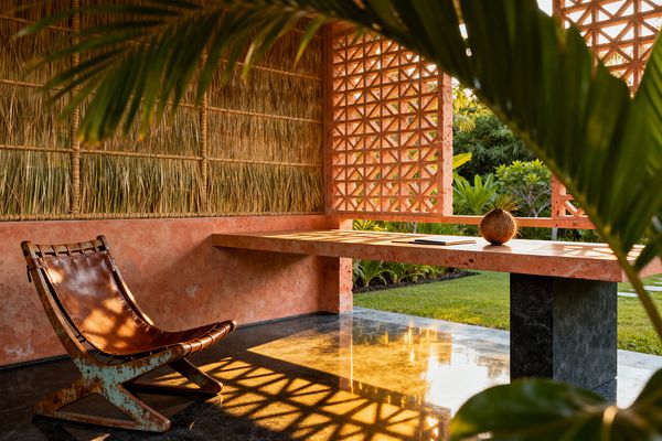 An open-air tropical home office with coral-pink plaster walls and a terracotta breeze-block screen opens to a lush garden. A cantilevered coral-toned stone desk on a dark stone pedestal sits beside a brown leather sling chair with a weathered frame. Woven palm-thatch wall panels and a polished charcoal floor catch warm sunlight, casting lattice shadows; a coconut and a slim notebook rest on the desk.