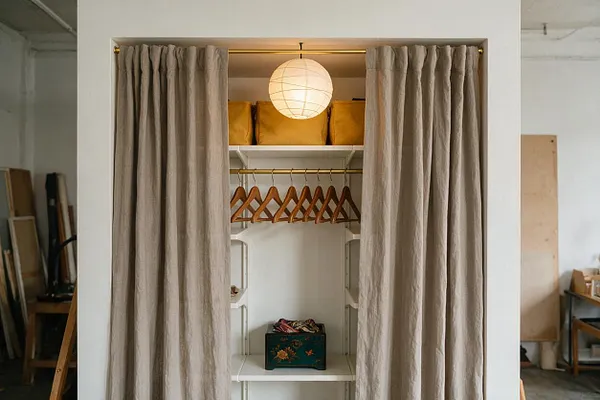 A small eclectic closet is framed by floor-length beige linen curtains and lit by a round paper pendant. Inside, white shelves hold mustard storage bins at the top, a row of wooden hangers on a brass rod, and a decorative floral box on the lower shelf.