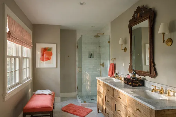 A master bathroom with a double wood vanity, marble countertop, and brass fixtures beneath an ornate dark wood mirror. A glass shower with white tile sits at the back, while a coral upholstered bench, matching bath mat, and pink window shade add color beside large windows.