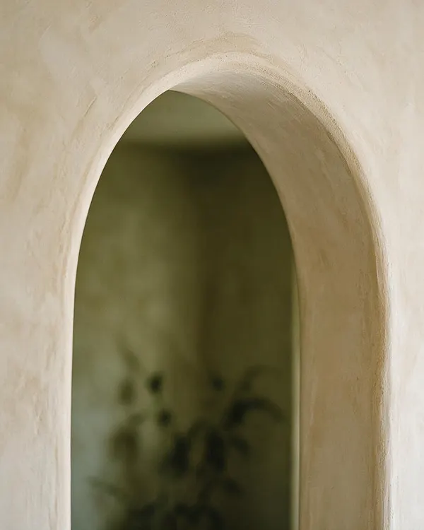 A close-up view of a smooth, rounded plaster arch with an off-white, textured finish. Beyond the opening, a softly blurred green interior suggests plants and a calm biophilic atmosphere.