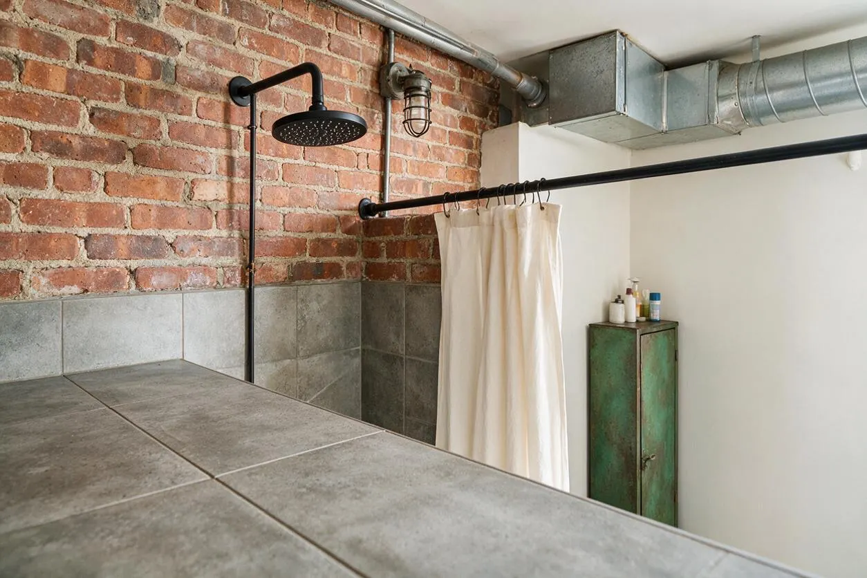A master bathroom with an industrial style featuring an exposed red brick wall, gray tile surround, black metal shower fixtures, and visible galvanized ductwork near the ceiling. A cream shower curtain hangs beside a weathered green metal cabinet, creating a raw yet refined loft-like atmosphere.