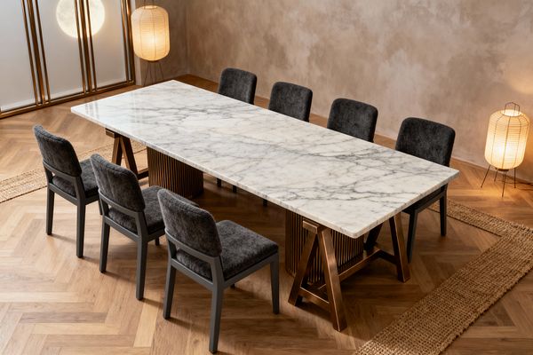 A contemporary dining room centers on a long, rectangular white marble table with pronounced gray veining, supported by a fluted cylindrical pedestal and trestle legs in warm bronze. Eight charcoal-upholstered chairs with dark frames sit on a herringbone oak floor bordered by jute runners, while two paper floor lanterns and a shoji-style screen with frosted panels cast soft, ambient light across taupe plaster walls, creating a serene, refined atmosphere.