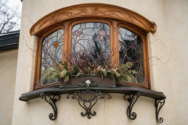 A cream-colored exterior wall features a rounded Art Nouveau bay window with flowing carved wood trim and stained-glass panels decorated with floral motifs. Below it, a curved dark metal shelf with ornate scroll brackets holds a planter filled with winter greenery and branches.