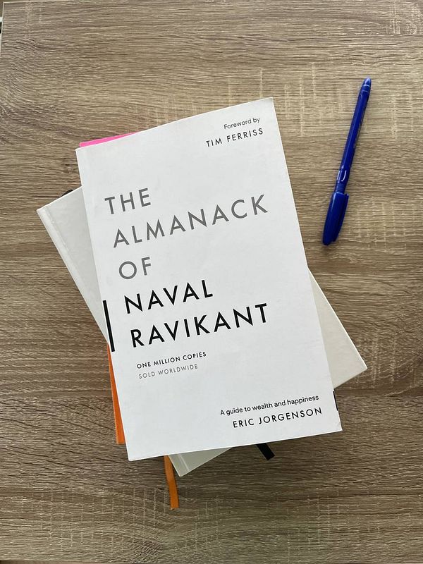 The image features a book titled 'The Almanack of Naval Ravikant' resting on a wooden surface.