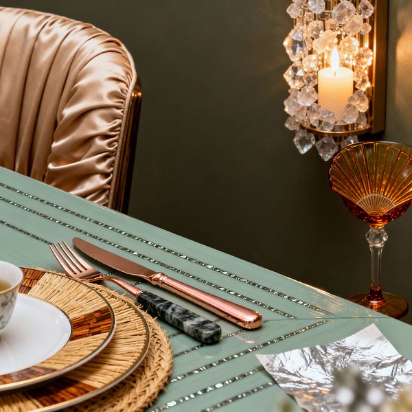 Close-up of a Vintage Glam dining scene featuring a mint green glossy table with slim metallic inlays and layered place settings: a rattan charger, wood-trimmed porcelain plate, and teacup. Rose-gold cutlery sits beside a spreader with a marbled black handle. An amber fluted coupe and a brass wall sconce draped in faceted crystals with a lit candle glow against a dark olive wall near a ruched champagne satin chair.