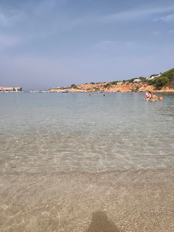 A young man sits on a rocky hill overlooking a serene beach scene with calm water, blue sky, and mountains in the background.
