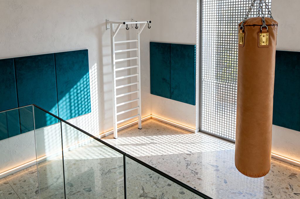 A bright modern home gym features a hanging tan leather punching bag with brass fittings, white Swedish wall bars with hooks, and deep teal upholstered wall panels mounted on white plaster walls. Sunlight filters through a perforated metal screen, casting dotted shadows across a pale terrazzo floor and a glass balustrade, while concealed baseboard LED strips create a warm glow that highlights the clean lines and minimal palette.