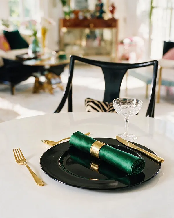 A stylish dining vignette features a glossy black plate topped with a rolled emerald-green napkin secured by a gold ring, set on a white table with gold flatware and a clear crystal glass. In the softly blurred background, black accent chairs, patterned pillows, and metallic furnishings reinforce a luxurious Hollywood Regency mood.