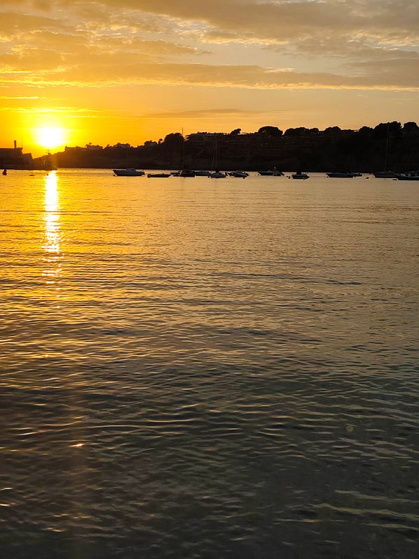 Sun setting over a calm body of water with boats anchored near the shore and silhouetted land in the distance.