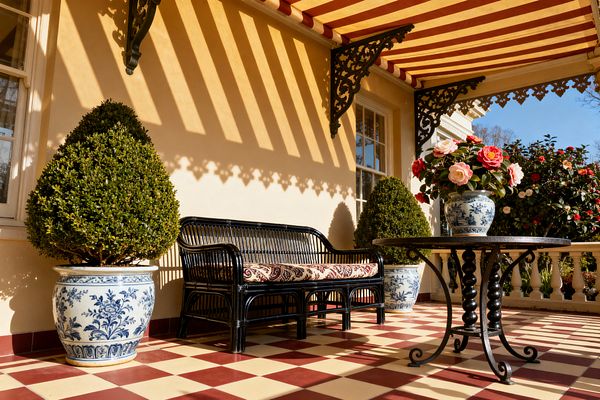 A Victorian-style patio with a red-and-cream striped awning casting patterned shadows over buttercream walls and ornate iron brackets. On a burgundy-and-ivory checkerboard tile floor, a black rattan settee with a damask cushion sits beside a round wrought-iron table holding a blue-and-white porcelain vase of pink and white flowers; matching planters contain clipped topiary, with mullioned windows and a white balustrade framing a flowering garden.