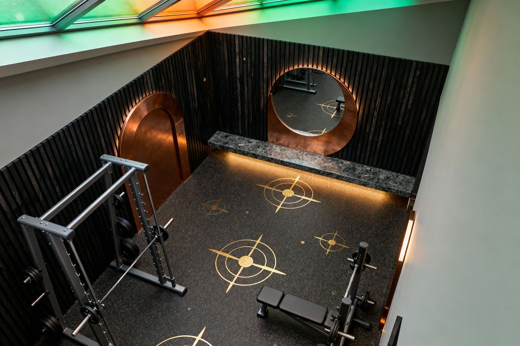 A moody home gym features black vertical wood-slat walls, an arched copper-clad doorway, and a large round mirror set within a copper panel, glowing with warm backlighting. The speckled black rubber floor shows gold circular compass-like inlays, a dark stone console stretches beneath the mirror, a silver power rack and weight bench occupy the space, and a sloped skylight with green-to-amber tinted panes washes the room in atmospheric color.