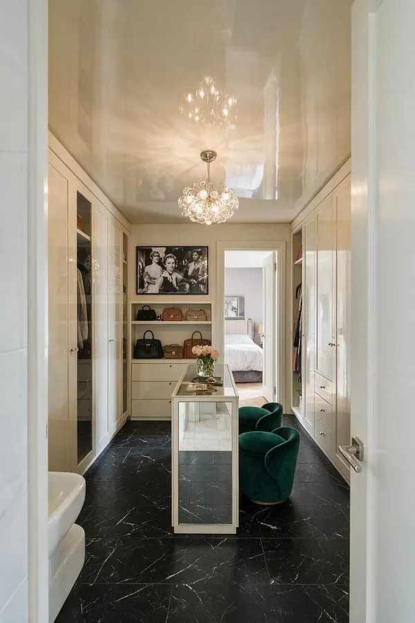 A narrow closet dressed in Hollywood Regency style features glossy cream built-in wardrobes, a mirrored center island, a crystal pendant chandelier, and black marble-look flooring. Velvet green stools, a framed black-and-white portrait, and neatly displayed handbags add glamour and sophistication.