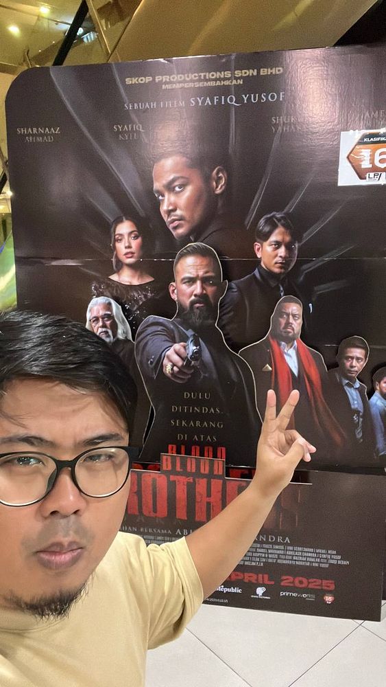 A person poses in front of a promotional poster for the movie 'Blood Brothers'.