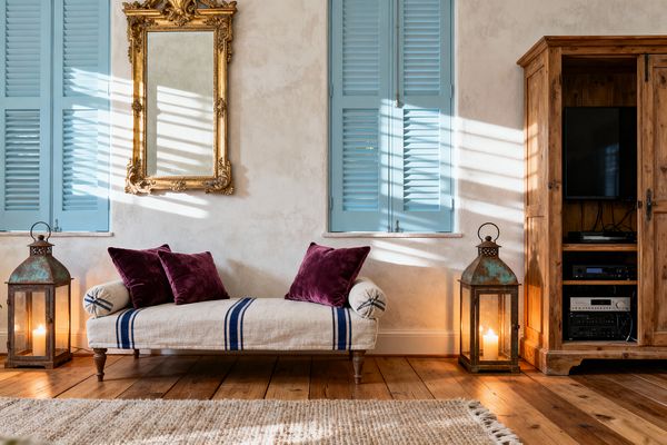 A cozy French Country media room shows a rustic pine armoire open to a TV and audio components, a French daybed upholstered in natural linen with navy stripes and plum velvet pillows, and two patinated metal-and-glass lanterns with candles. Sky-blue shutters cast slatted sunlight across textured ivory plaster walls beneath an ornate gilded mirror, with wide-plank wood floors and a nubby neutral rug grounding the scene.
